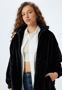 Sort fleecejakke med høj krave, lynlåslukning og oversized pasform, båret over en hvid skjorte med knapper og en cropped top.