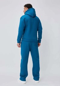 Sweat à capuche bleu sarcelle et joggings assortis en tissu doux et texturé. Comprend une poche kangourou et des poignets côtelés. Baskets blanches visibles.
