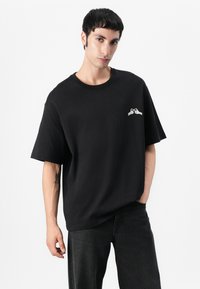 Black cotton t-shirt with short sleeves, featuring a small white graphic on the left chest. Plain design and relaxed fit.