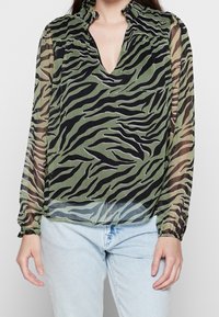 Blusa verde trasparente con un motivo a strisce tigre in nero e grigio, scollatura a V e polsini elasticizzati. Abbinata a jeans azzurri chiari.