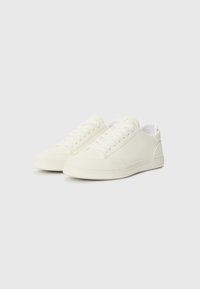 White sneakers with textured fabric, rounded toes, and flat rubber soles. Features lace-up closure and minimalistic design.