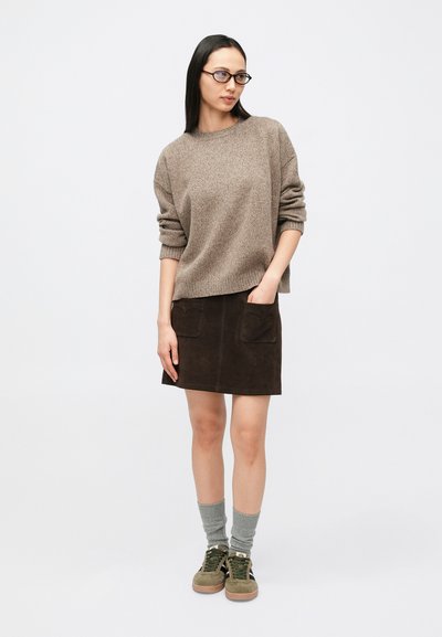Femme portant un pull marron, une jupe marron foncé avec poches, des chaussettes grises et des baskets vertes, se tenant devant un fond clair uni.