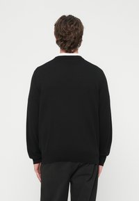 Homme aux cheveux bouclés bruns portant un pull noir et un pantalon noir, de dos sur un fond clair uni.
