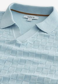 Jasnoniebieska dziergana koszulka polo z teksturowanym wzorem w kratę, dekoltem w kształcie V oraz kontrastowym kołnierzykiem z złotym akcentem.