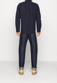 Camicia blu navy a maniche lunghe con polsini abbottonati e orlo arrotondato, abbinata a pantaloni in denim scuri dal fit morbido, arrotolati sui polsini, e scarpe color sabbia.