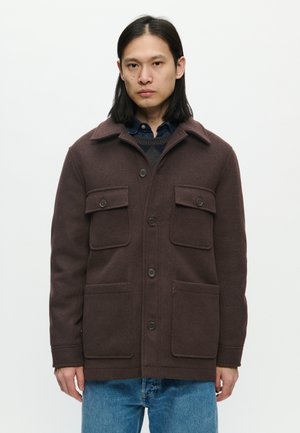 Homme aux longs cheveux noirs portant une veste marron foncé boutonnée avec quatre poches avant et un jean bleu sur un fond uni.