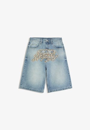 Blaue Jeans mit großer stilisierter Stickerei und kleinen Strasssteinen im vorderen Oberschenkelbereich, kombiniert mit einem weißen Hemd.