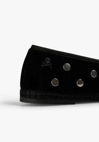 Mocasines de terciopelo negro con tachuelas redondas plateadas y un sutil logotipo de calavera y tibias cerca del talón, sobre un fondo blanco.