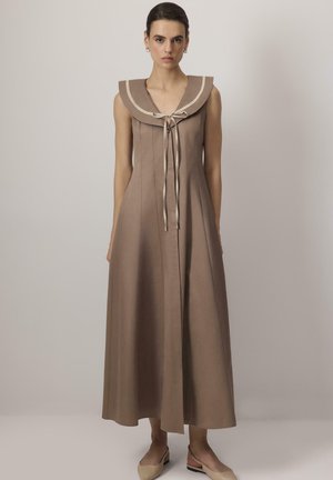 Ženska stoji in nosi brezrokavno rjavo midi obleko s širokim ovratnikom in pentljo, v kombinaciji z bež nizkimi čevlji.