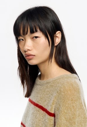 Suéter de punto en beige con rayas rojas, con un escote redondo y una tela suave y texturizada. La modelo tiene el pelo largo y oscuro y lleva pendientes mínimos.