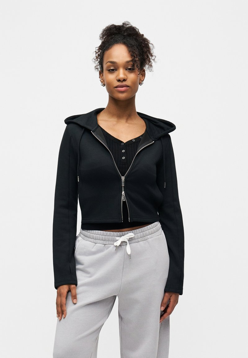 Jeune femme aux cheveux bouclés portant un sweat à capuche court noir zippé par-dessus un haut noir à boutons et un pantalon de survêtement gris clair à cordon de serrage.