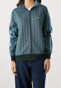 Grön zip-up jacka med geometriskt mönster, ribbstickade muddar och fåll, detaljerade sidoseam med blå ränder och en liten logotyp på bröstet.