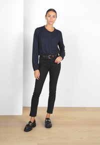 Blusa azul marino de botones con mangas largas, combinada con jeans skinny negros y mocasines negros de plataforma, sobre un fondo claro.
