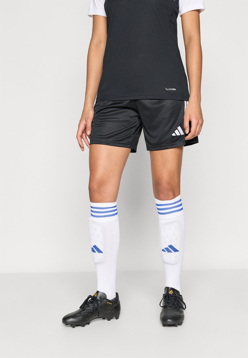 Μαύρο αθλητικό σορτς με λευκό και μαύρο μπλουζάκι τύπου φανέλας, συνδυασμένο με λευκές κάλτσες με μπλε ρίγες και μαύρα παπούτσια ποδοσφαίρου.