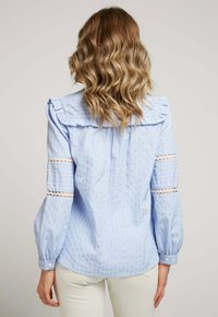 Blouse rayée bleu et blanc avec un col volanté, des manches bouffantes et des détails en dentelle sur les bras. Fermeture à boutons aux poignets. Tissu à texture lisse.