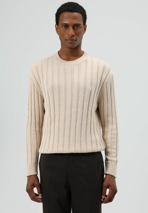 Beige geribbelde trui met een ronde hals en lange mouwen, voorzien van een geprint patroon en nauwsluitende manchetten. Gecombineerd met donkere broek.