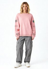 Svijetloružičasti dukserica s grafikom na rukavima, u kombinaciji s slobodnim sivim cargo hlačama. Dukserica ima rebrasti izrez i donji rub. Bijele tenisice.