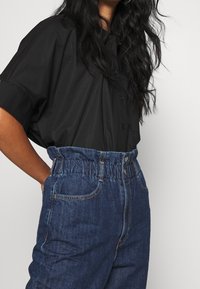 Svart kortärmad skjorta med knappar, kombinerad med högmidjade blå denimjeans med elastisk midja och synliga sömmar.