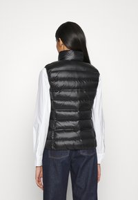 Czarna, bezrękawnikowa kamizelka puchowa z wysokim kołnierzem, błyszczącą fakturą i poziomym pikowaniem. Noszona na białej koszuli i dżinsach.