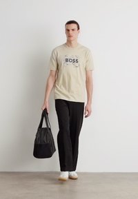 Beige bavlněné tričko s potiskem "BOSS", černé kalhoty a bílé tenisky, nese černou látkovou tašku. Minimalistické pozadí.