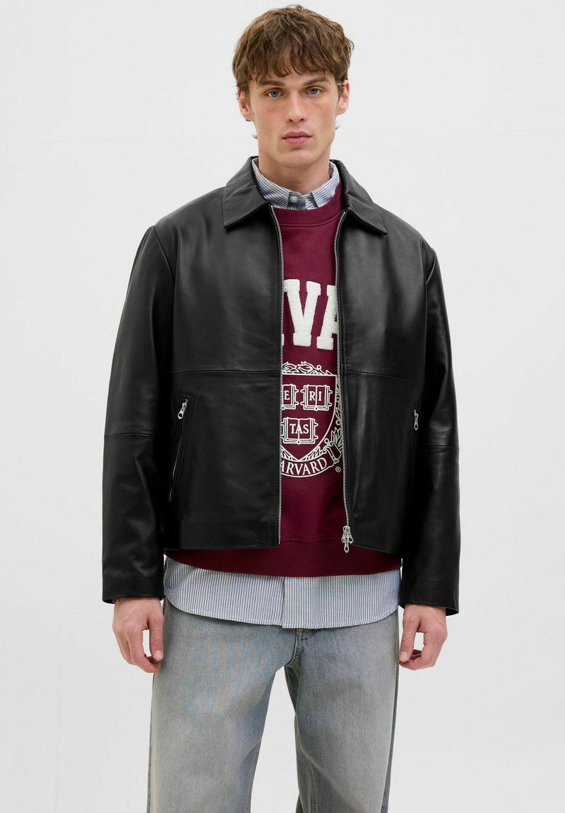 Schwarze Lederjacke mit durchgehendem Reißverschluss, mit einem Kragen-Halsbereich und zwei seitlichen Taschen. Über einem bordeauxfarbenen Sweatshirt getragen.