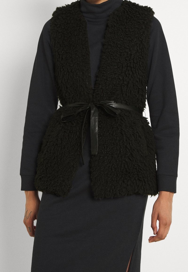 Personne portant un gilet sans manches noir en matière duveteuse, attaché avec une ceinture en cuir noir, par-dessus une robe sombre à manches longues avec une fente sur le côté.