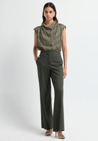 Femme debout portant un haut sans manches à motifs écossais noir, beige et jaune, un pantalon large gris foncé taille haute et des talons beiges.