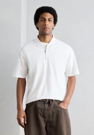 Młody mężczyzna z krótkimi kręconymi włosami, ubrany w biały, krótkawy sweter z zamkiem i brązowe spodnie, stojący z jedną ręką w kieszeni.