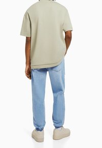 Person som bär en beige, oversized kortärmad skjorta, ljusblå denimjoggingbyxor och beige sneakers, står med ena handen i fickan.