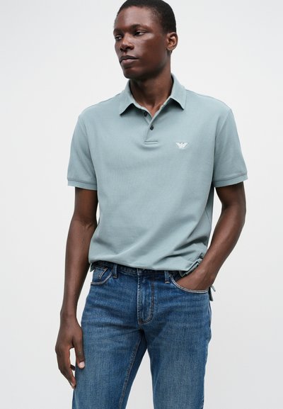 Ανοιχτό πράσινο πόλο μπλουζάκι με γιακά και τρία κουμπιά, που διαθέτει μικρό λογότυπο αετού. Φορεμένο με μπλε τζιν παντελόνι. Λείος Texture.