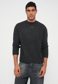 Άνδρας με σκούρο γκρι μάλλινο πουλόβερ μακρυμάνικο με μικρό λογότυπο κοντά στον γιακά και ξεθωριασμένο μαύρο τζιν, στέκεται μπροστά σε απλό λευκό φόντο.