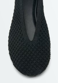 Chaussure ballerine plate en maille noire avec un motif texturé et une ouverture en forme de V sur le dessus.