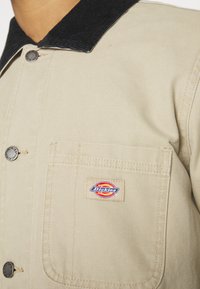 Beige Arbeitsjacke mit schwarzem Cordkragen, silbernen Knöpfen und gesticktem Dickies-Logo auf der Brusttasche. Robustes Material und klassisches Design.