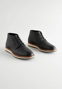Botas de tobillo de cuero negro con puntera redondeada, diseño de cordones y suela de goma blanca contrastante. Parte superior texturizada y detalles de costura en marrón.