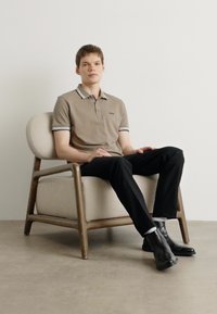 Bež majica polo s črnim obrobom, črne hlače in črne škornje nosi model, ki sedi v svetlo obarvanem lesenem stolu.