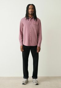 Langärmliges, knopfbares Hemd in Mauve mit einer Brusttasche, kombiniert mit schwarzen Hosen und grauen Turnschuhen, im Stehen vor einem neutralen Hintergrund.