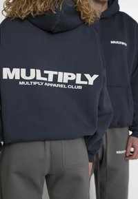 Felpa con cappuccio blu navy con logo "MULTIPLY" bianco e scritta "MULTIPLY APPAREL CLUB" sul retro. Pantaloni della tuta grigi con dettaglio tasca posteriore.