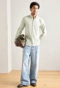 Ανοιχτοπράσινο πουκάμισο με κουμπιά και μακριά μανίκια, συνδυασμένο με φαρδιά ανοιχτό μπλε τζιν και καφέ παπούτσια. Κρατάει τσάντα καφέ και ελιάς.