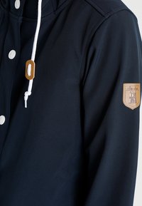 Navy windbreaker jacket with a smooth, waterproof fabric; features white drawstring, silver snap buttons, and a textured tan patch on the sleeve.