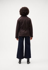 Giacca a maniche lunghe marrone scuro con cintola, abbinata a jeans in denim scuro a gamba larga e stivali neri con tacco, vista da dietro.