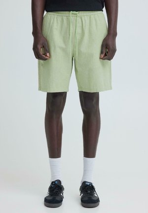 Mężczyzna w jasnych zielonych szortach ze ściągaczem, czarnych tenisówkach i białych skarpetach za kostkę, stojący na tle jednolitego jasnego tła.