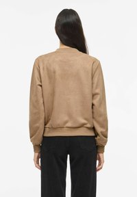 Pruun seemisnahast pommitaja-stiilis jakk pikkade varrukatega, soonikkoelsete mansettide ja vöökohaga, sileda tekstuuriga ning vabalt langeva lõikega. Mustad püksid sobituvad selle alla.