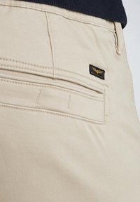 Beige bomuldsbukser med en baglomme med et sort logo-tag og subtil syning, der fremhæver en glat tekstur og skræddersyet design.