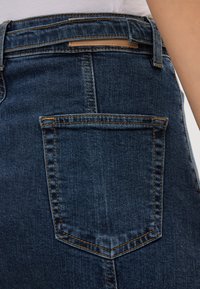 Jeans in denim scuro presentano una tasca posteriore con cucitura arancione a contrasto e un piccolo dettaglio in pelle vicino alla vita. Texture liscia osservata.
