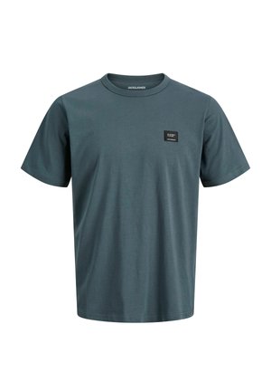 Garçon debout portant une casquette noire, une veste zippée noire, une chemise bleue, un pantalon ample foncé et des baskets noires et blanches sur un fond uni.