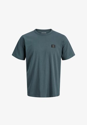 Dreng stående iført en sort kasket, sort lynlåstrøje, blå skjorte, løse mørke bukser og sorte og hvide sneakers mod en ensfarvet baggrund.