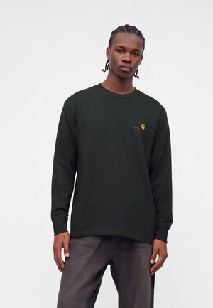 Giovane uomo con capelli intrecciati che indossa una maglia nera a maniche lunghe e pantaloni scuri su uno sfondo bianco uniforme.