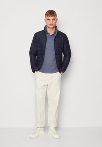 Giacca imbottita blu navy, colletto grigio, indossata sopra un polo azzurro chiaro, abbinata a pantaloni crema e sneaker bianche, in piedi contro uno sfondo semplice.