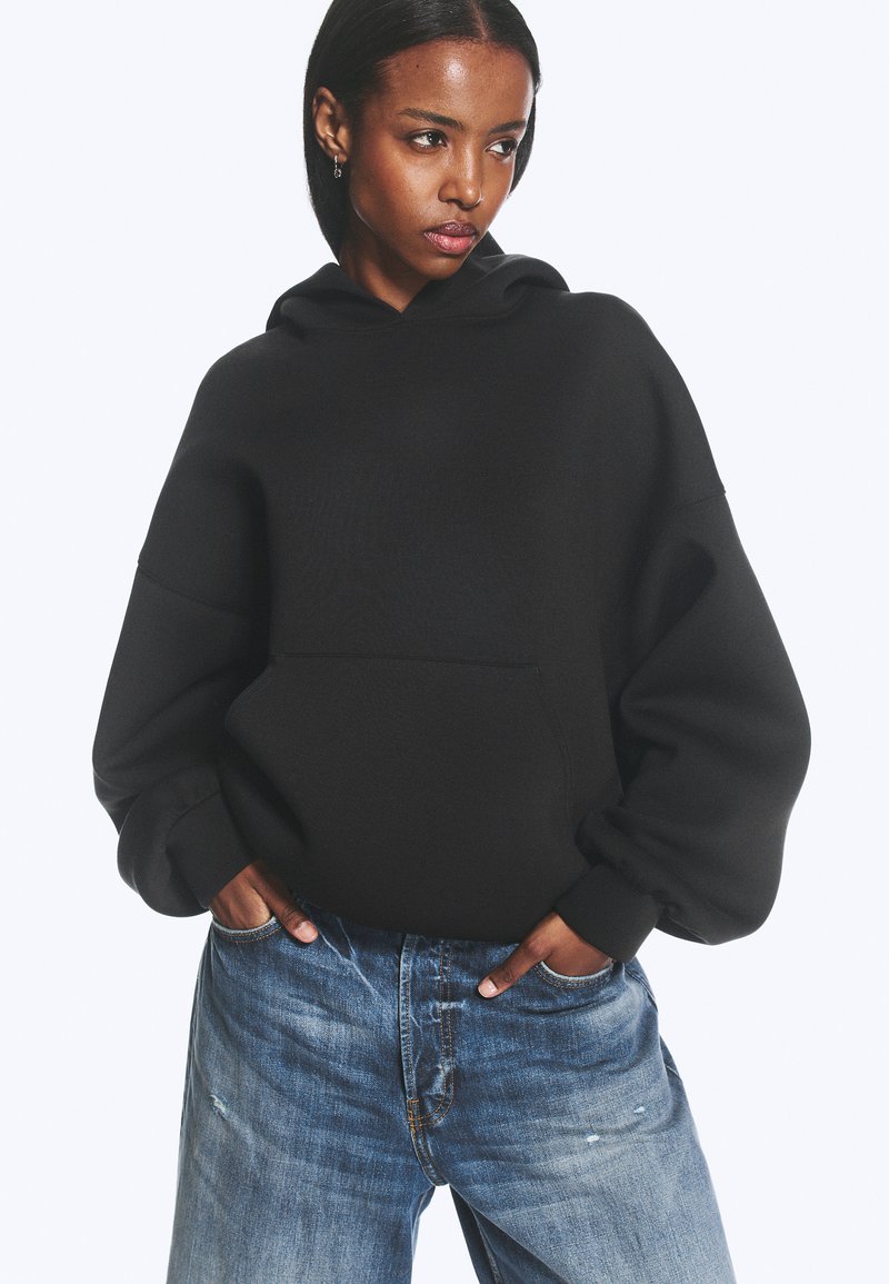 Kvinde med mørkt hår iført en sort oversized hættetrøje og blå slidte jeans, hænderne i lommerne, kigger til siden.