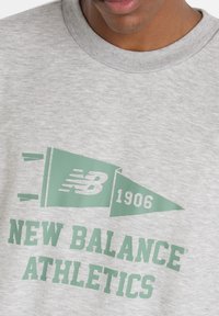 Ljusgrå sweatshirt med en mintgrön "New Balance Athletics"-logotyp och en triangulär fan-design med "1906"-detalj.